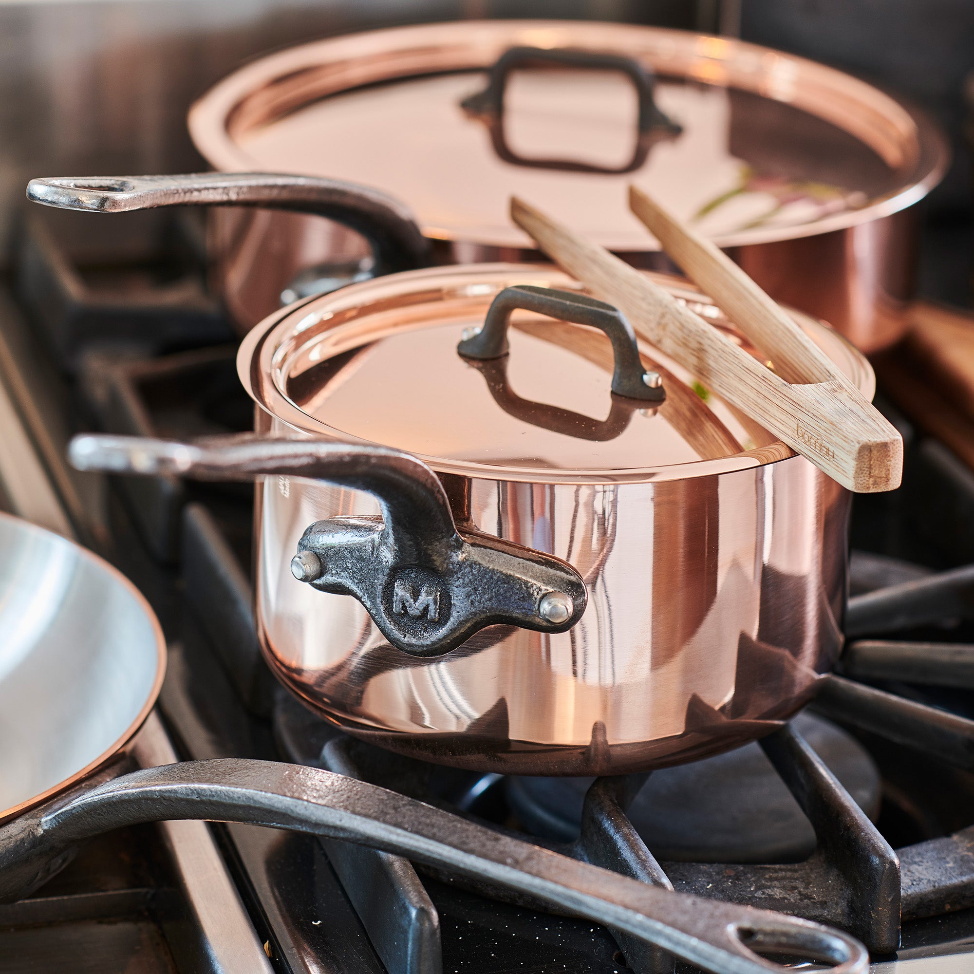 Mauviel M'Heritage M'150 CI copper frying pan set with two pans and cast iron handles, shown on a kitchen countertop.